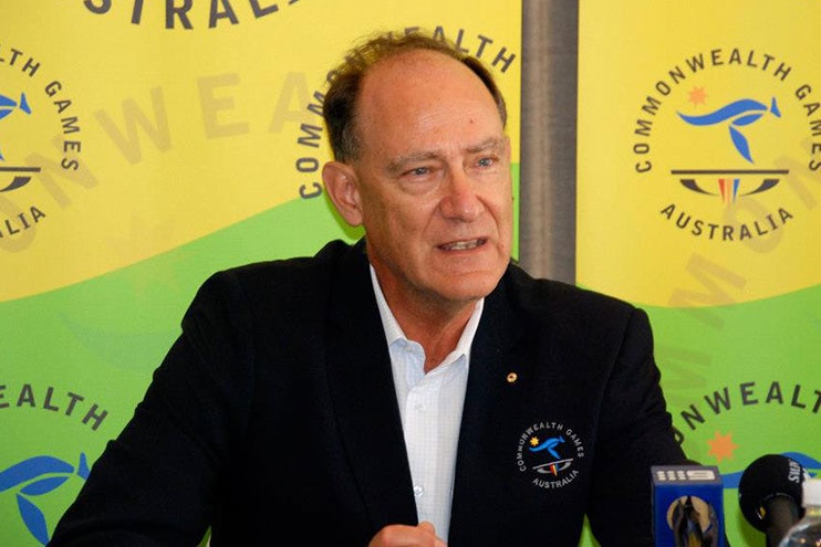 A man with a receding hairline, wearing a black  blazer, sits behind microphones in front of a Commonwealth Games backdrop.