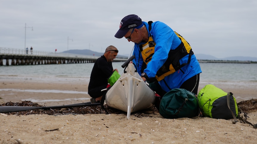 Two men packing goods into a kayak on the shore.