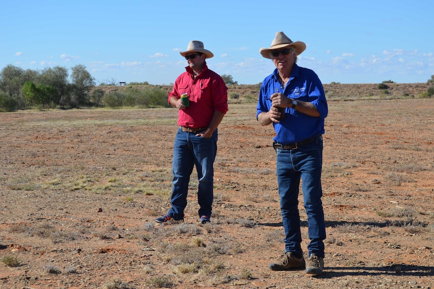 Two men in country attire and hats, with beet in there hands, standing on a rocky cricket pitch under a blue sky