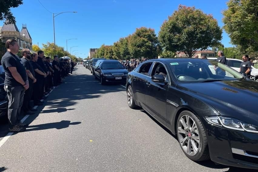 A procession of black cars moves down a street, flanked by a guard of honour.