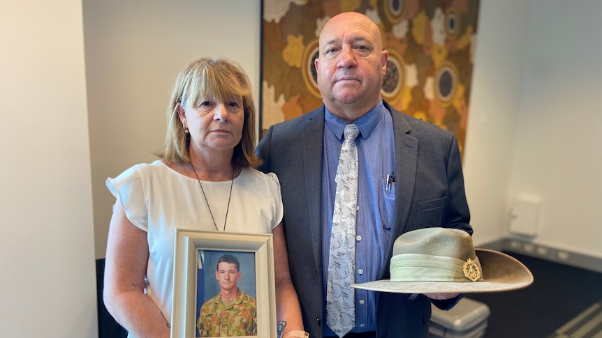 An elderly couple stand holding a photo of their digger son and slouch hat.