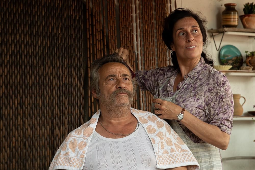 A standing woman cuts the hair of a seated man as both look at something out of shot.