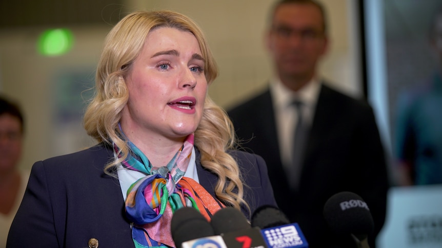 A woman with blonde hair and a colourful scarf answers a question standing behind microphones