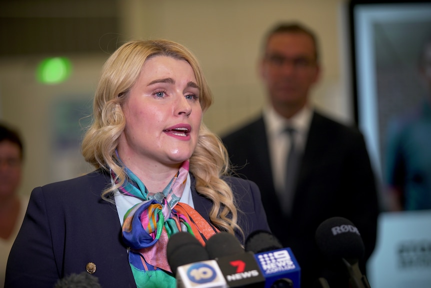 A woman with blonde hair and a colourful scarf answers a question standing behind microphones