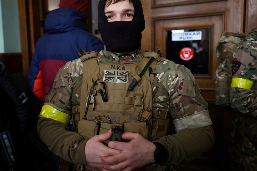 A young man in combat fatigues looks at the camera through a balaclava, he has a British patch on.