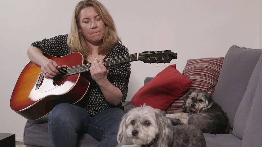 Musician Rebecca Brown plays guitar on the couch next to her two dogs.