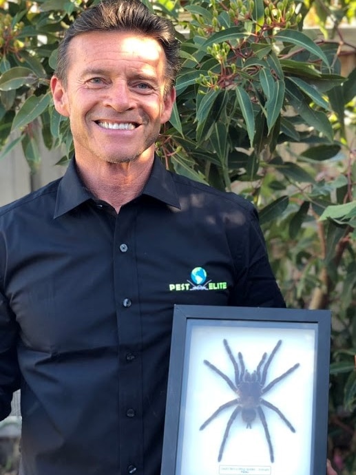 A smiling man in a dark button-up shirt holds a frame containing a gigantic spider.