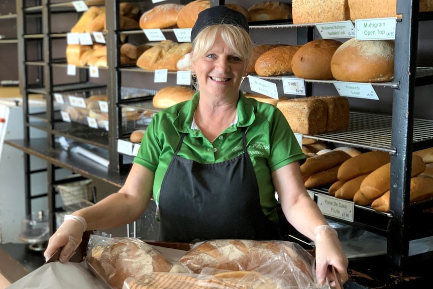 Ms Mayoss smiles and holds a pan of loaves of breads, in front of a rack of loaves.