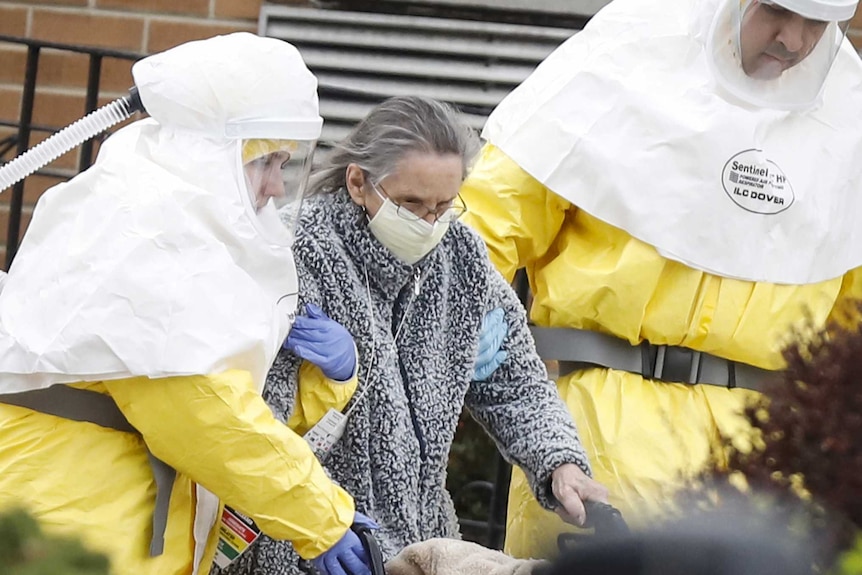 An elderly woman is assisted by medical officials in full protective suits who walk her out of a building