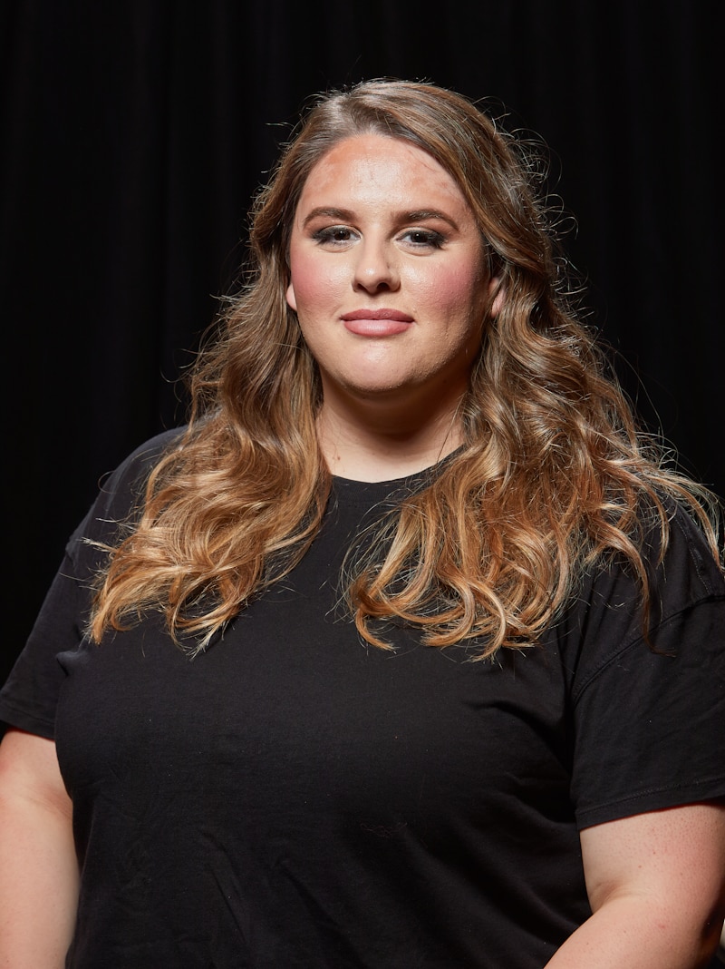 Plus-sized woman with brown wavy hair wearing dark tee shirt, against black background.