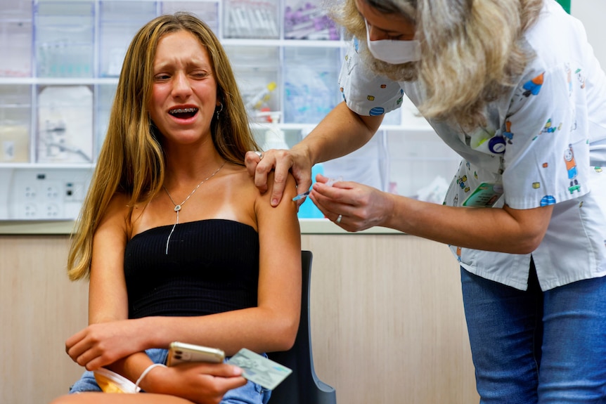 A teen girl with braces grimaces as a nurse injects her shoulder with a needle 