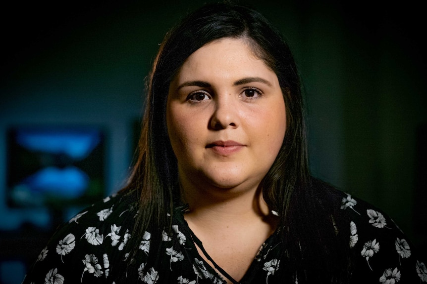 Renee Olofsson, with black hair wearing a dark blouse, stares into the camera with screens behind her.