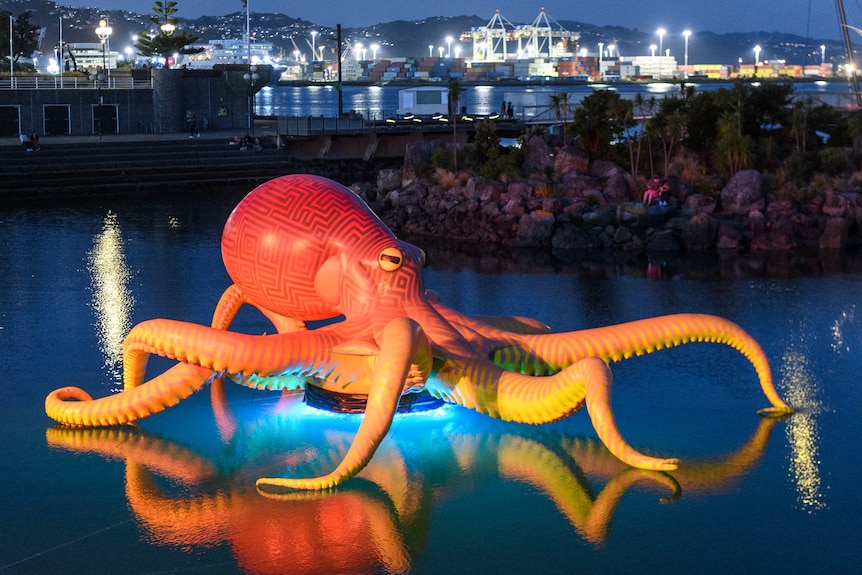 A giant orange octopus sits on a lit platform in the water with city lights in the background.
