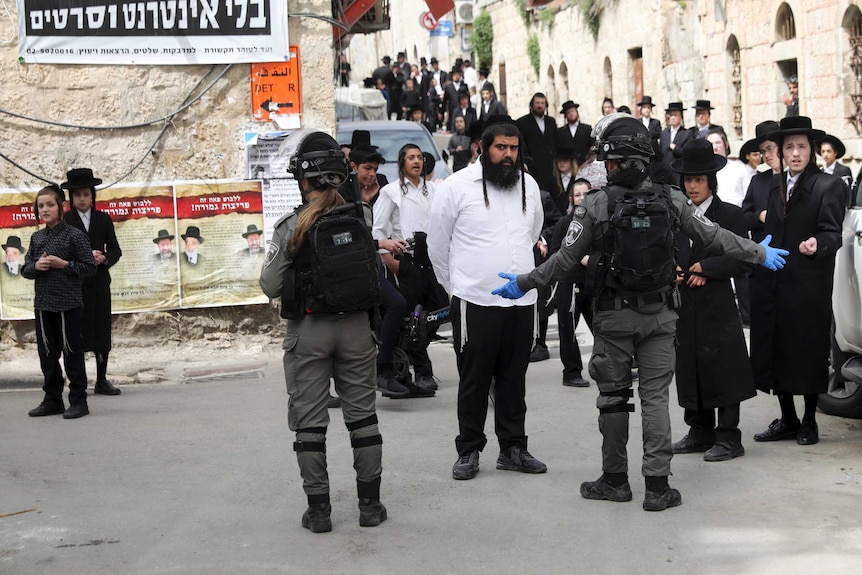 Police in black uniforms control a large group of men dressed in  orthodox Jewish clothing.