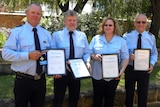 Four people in uniform stand in a garden holding certificates and a medal.