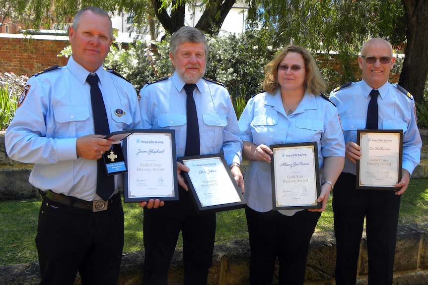 Four people in uniform stand in a garden holding certificates and a medal.