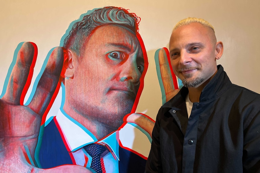 White man with blonde hair and black coat stands with portrait of Taika Waititi, a grey-haired man in a navy suit in 3D style