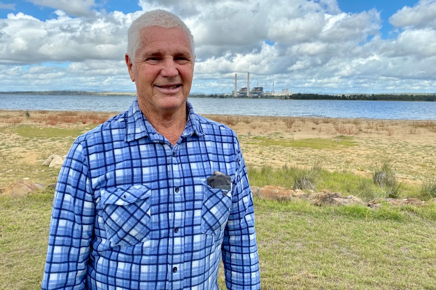 A man stands in front of a lake with a power station in the background.
