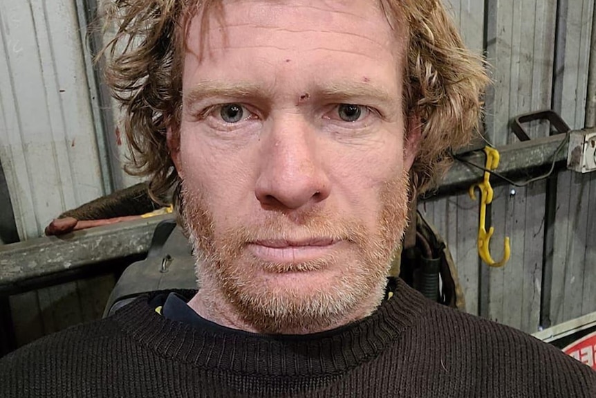 A middle aged white man with red stubble has a neutral facial expression.