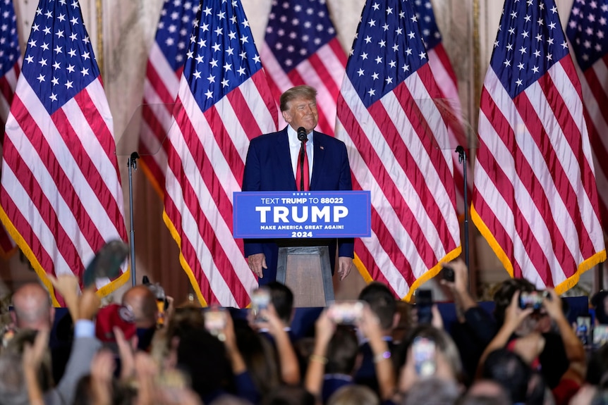 Donald Trump stands in front of a crowd with American flags behind him