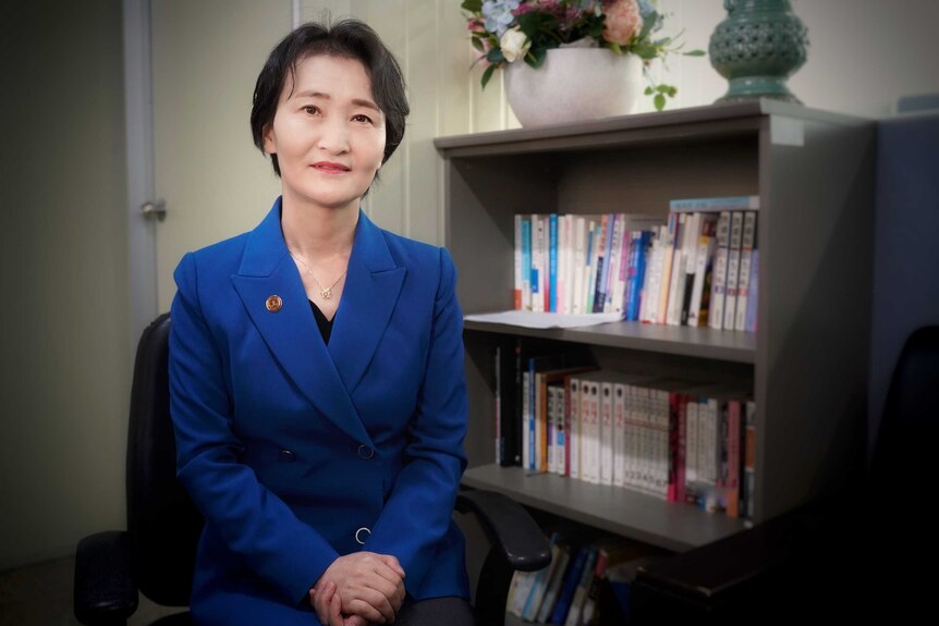 A Korean woman in a royal blue suit sitting next to a book case