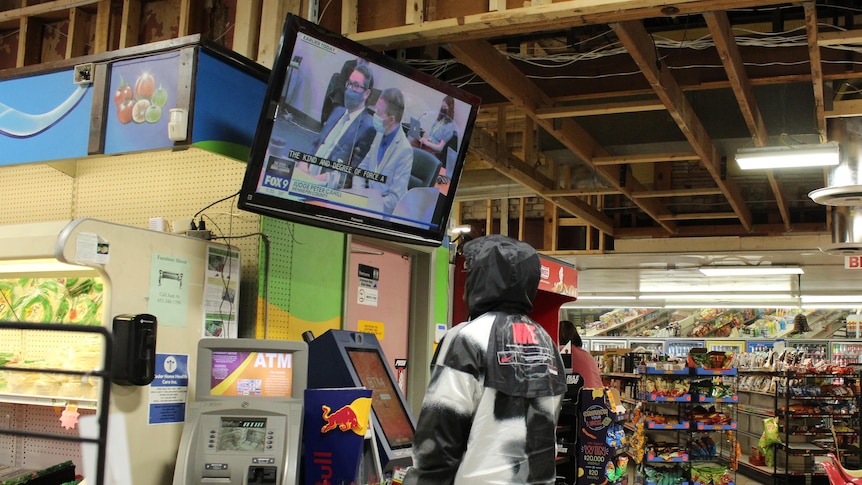 A man stands in a convenience store, watching a trial on TV 