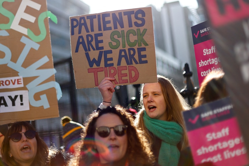 A group of nurses striking holding placards, one in focus reads "patients are sick we are tired" 