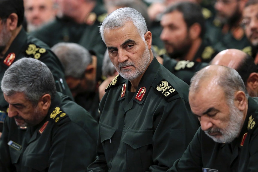 A man in military uniform with white hair glances at someone as he is surrounded by other men in uniform.