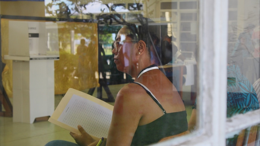 A woman sitting behind a glass window with a ballot paper in hand