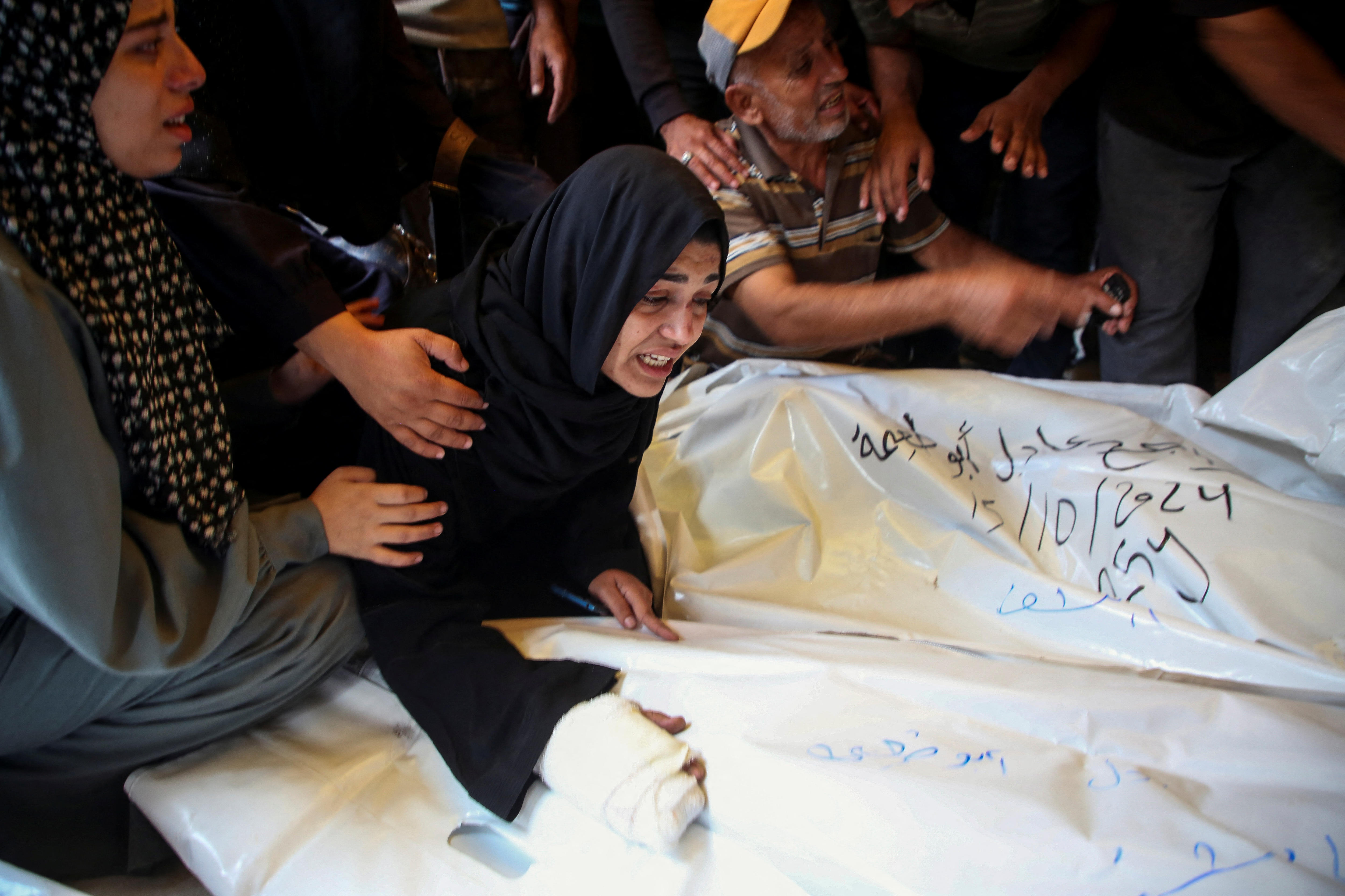 A woman in a black hijab cries as she reaches out to a white bodybag tagged in Arabic