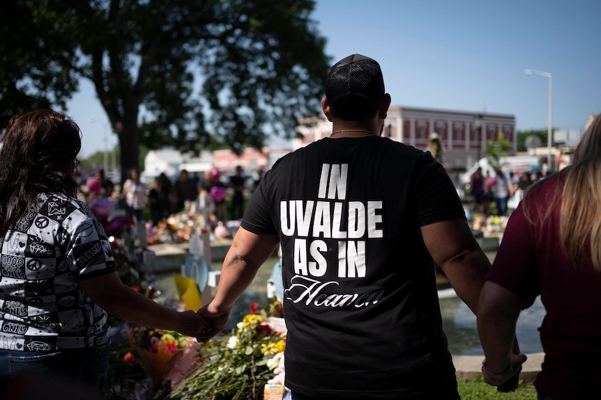 Pastor wearing a t-shirt that says "In Uvalde As In Heaven," leads a prayer circle at a memorial site