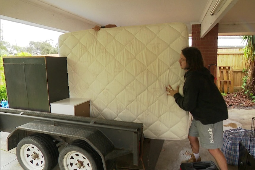 Corey loads a mattress onto a trailer with other furniture in the carport of his home
