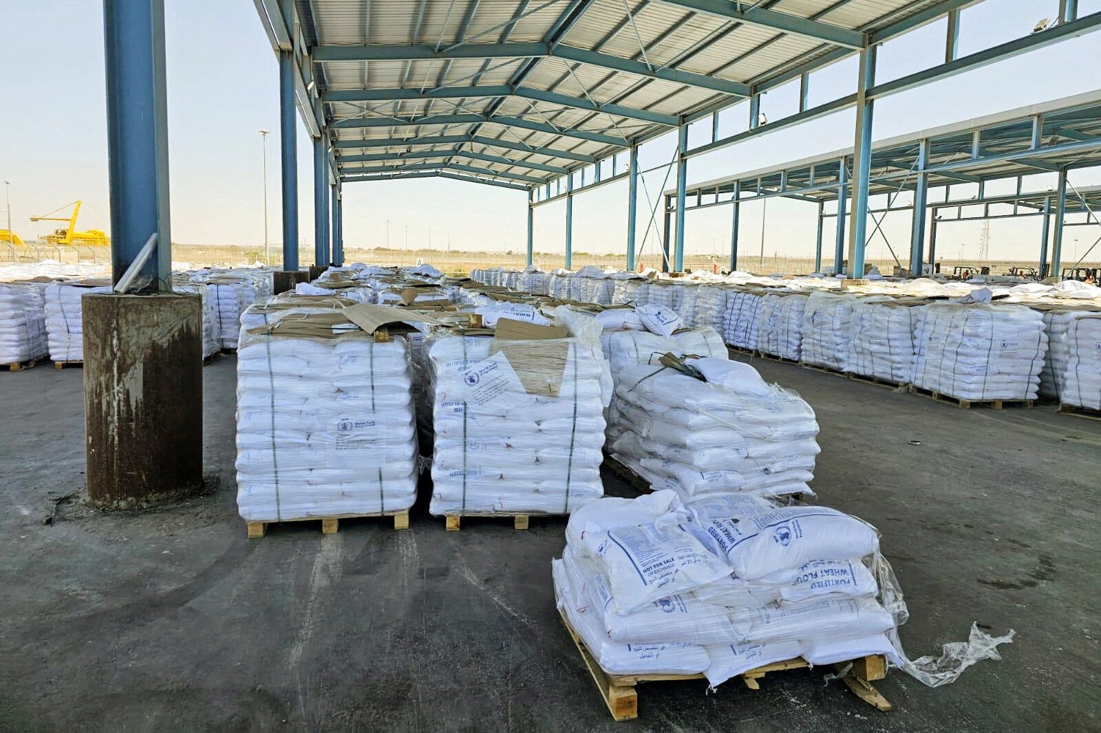 Multiple pallets of flour sacks sitting under an open air warehouse with dry hills seen in the background.