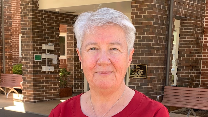 Viv Allanson wearing a red shirt with a logo on the breast and a name tag stands in front of a brick building