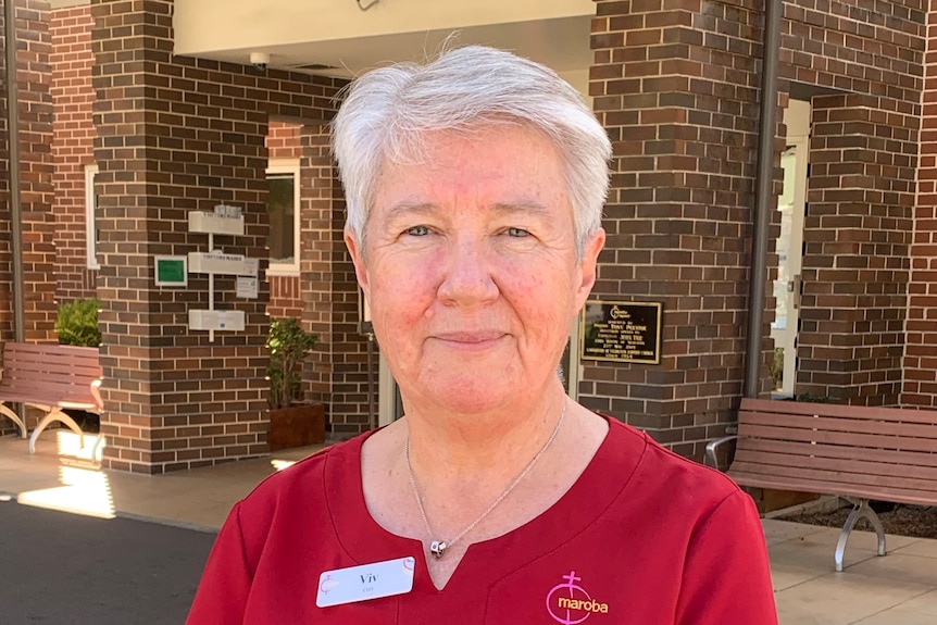 Viv Allanson wearing a red shirt with a logo on the breast and a name tag stands in front of a brick building