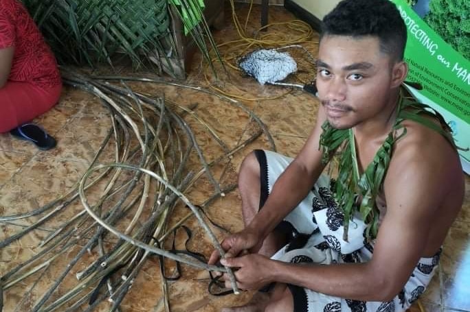 A man sits on the ground wooden sticks he is using make nets with.