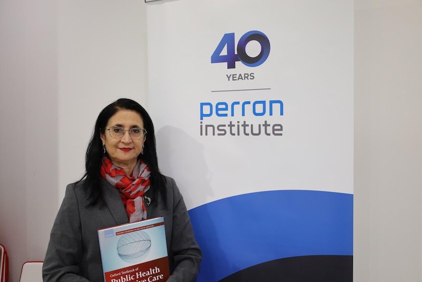 A woman with glasses holding a book in front of a banner with Perron Institute written on it.