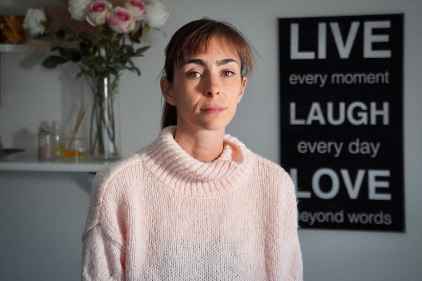 Sarah Kay sits in front of a vase of flowers and a "Live, Laugh, Love" poster, looking into the camera seriously.
