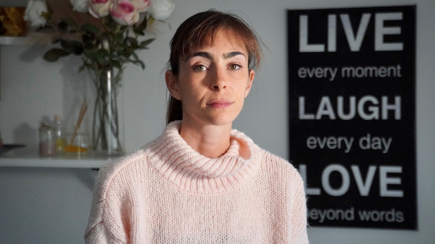 Sarah Kay sits in front of a vase of flowers and a "Live, Laugh, Love" poster, looking into the camera seriously.