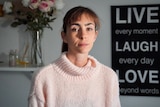 Sarah Kay sits in front of a vase of flowers and a "Live, Laugh, Love" poster, looking into the camera seriously.