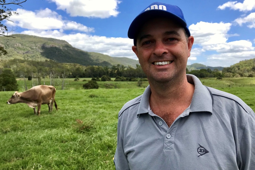 A smiling man in a cap stands in a lush green field, featuring a cow, beneath a mostly clear sky.