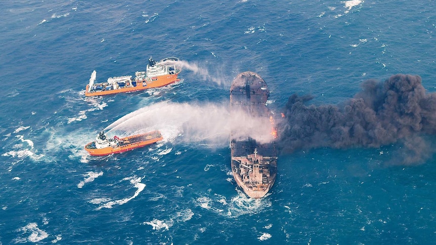 Two ships spray water onto a tanker that is ablaze and billowing black smoke in the middle of the ocean