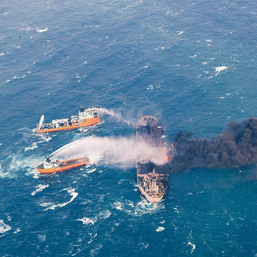 Two ships spray water onto a tanker that is ablaze and billowing black smoke in the middle of the ocean