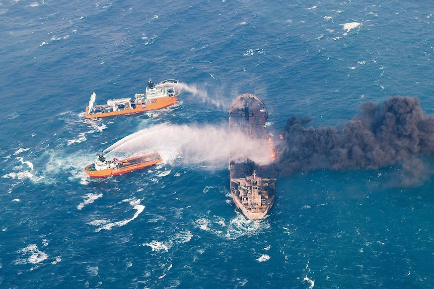 Two ships spray water onto a tanker that is ablaze and billowing black smoke in the middle of the ocean