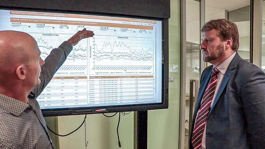 Two Western Power employees stand beside a big TV screen looking at graphs.