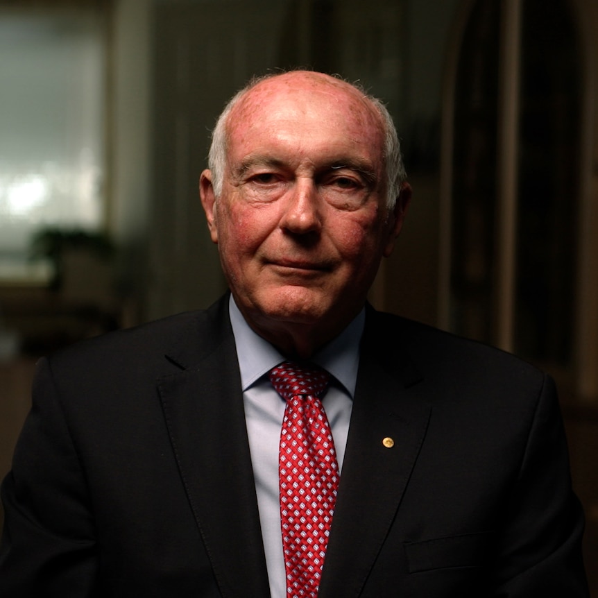 Warren Truss sits in an office, dressed in a suit and red tie, looking into camera with a serious expression.