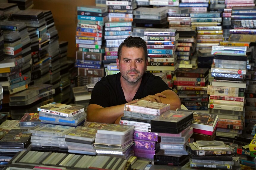 A man wearing a black t-shirt stands amongst a pile of colourful video tapes