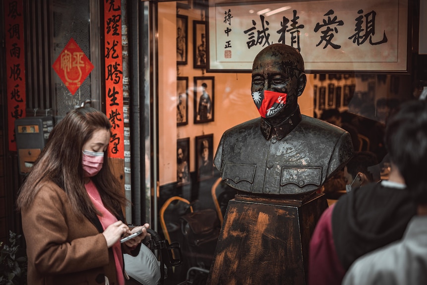 A masked woman looks at a bronze statue wearing a mask made with Taiwan's flag