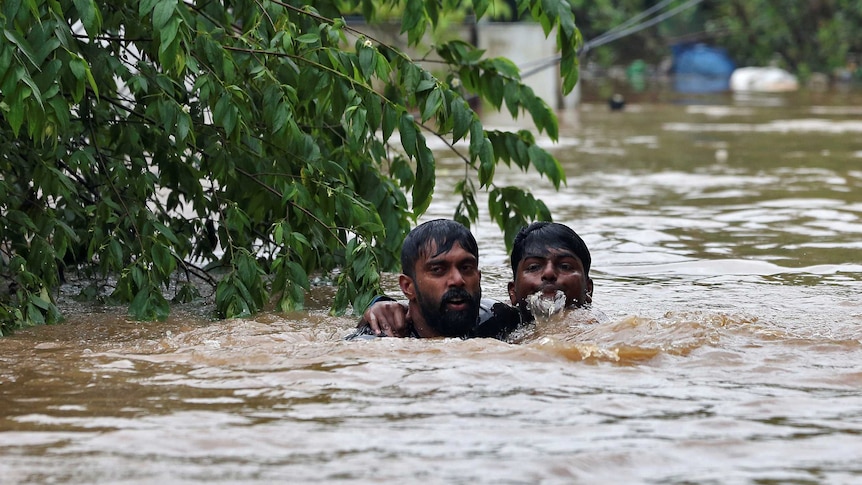 Two men walk in flooded waters. One man has one arm around another man who is helping him stay afloat.