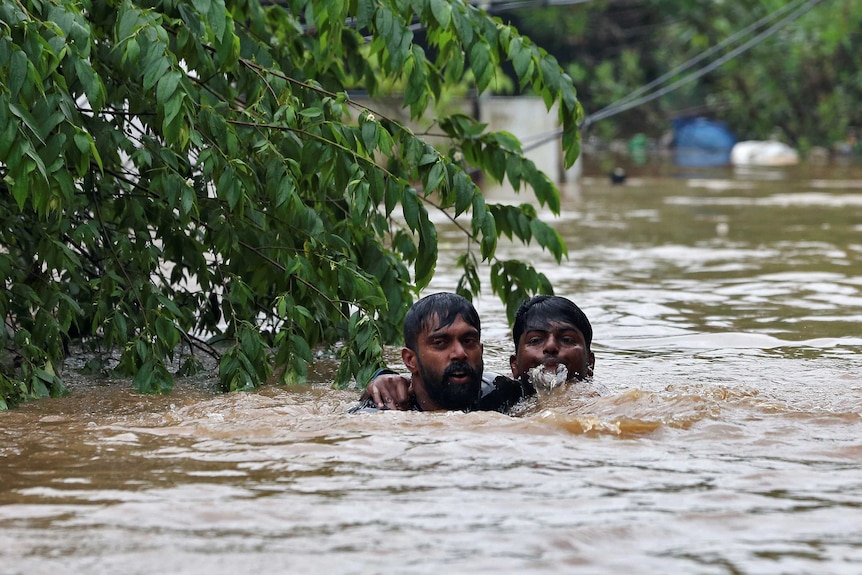 Two men walk in flooded waters. One man has one arm around another man who is helping him stay afloat.